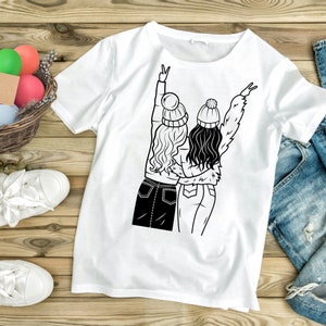 May include: White t-shirt with a black line drawing of two women wearing winter hats and jeans, standing back to back with their arms around each other and making peace signs with their hands.