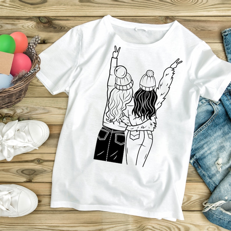 May include: White t-shirt with a black line drawing of two women wearing winter hats and jeans, standing back to back with their arms around each other and making peace signs with their hands.