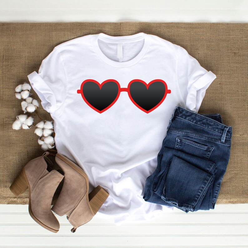 May include: White t-shirt featuring a graphic of heart-shaped sunglasses with black lenses and red frames. The shirt is paired with blue denim jeans and beige ankle boots. The items are arranged on a neutral-toned surface.