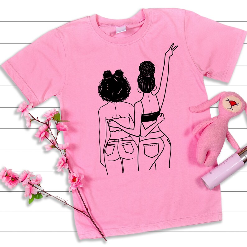 May include: Pink t-shirt with a black line drawing of two women with their arms around each other. The women are wearing jeans and have their hair pulled back.