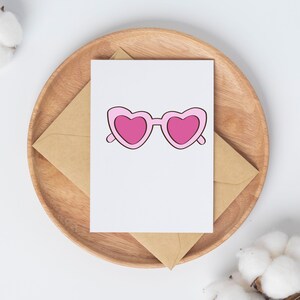 May include: A greeting card featuring a pink heart-shaped sunglasses design. The card is placed on a brown envelope, resting on a wooden plate. The background is white, with cotton buds in the corners.
