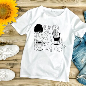 May include: A white t-shirt with a black line drawing of three women standing together. The women are wearing different hairstyles and outfits. The design is centered on the shirt.