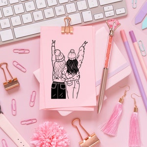 May include: A pink card with a black line drawing of two friends wearing winter hats and holding hands. The card is on a pink surface with other craft supplies.