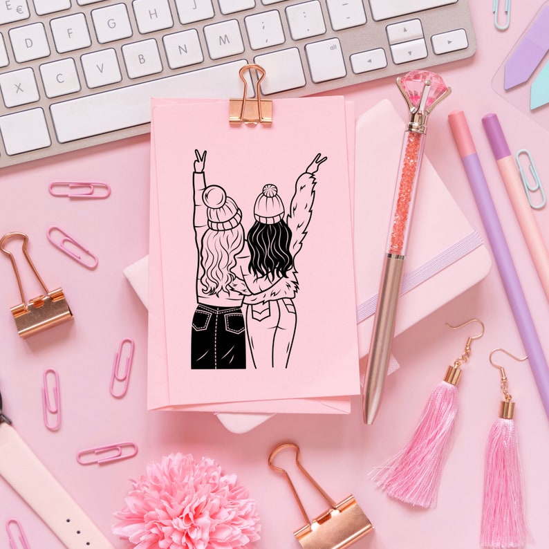 May include: A pink card with a black line drawing of two friends wearing winter hats and holding hands. The card is on a pink surface with other craft supplies.