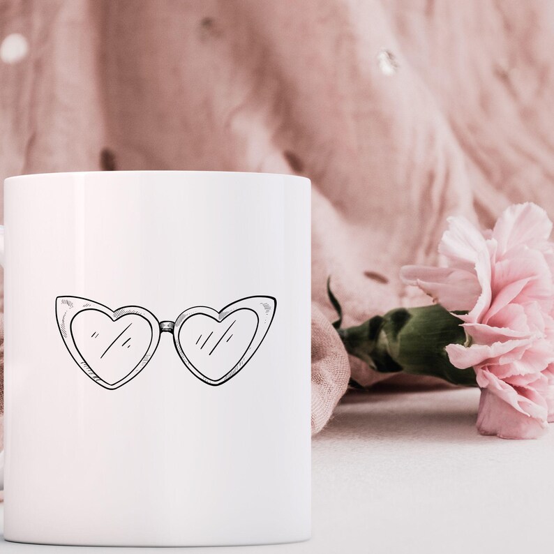 May include: White ceramic mug featuring a black line drawing of heart-shaped sunglasses. The mug is set against a soft pink background with a pink flower, creating a feminine and stylish aesthetic.