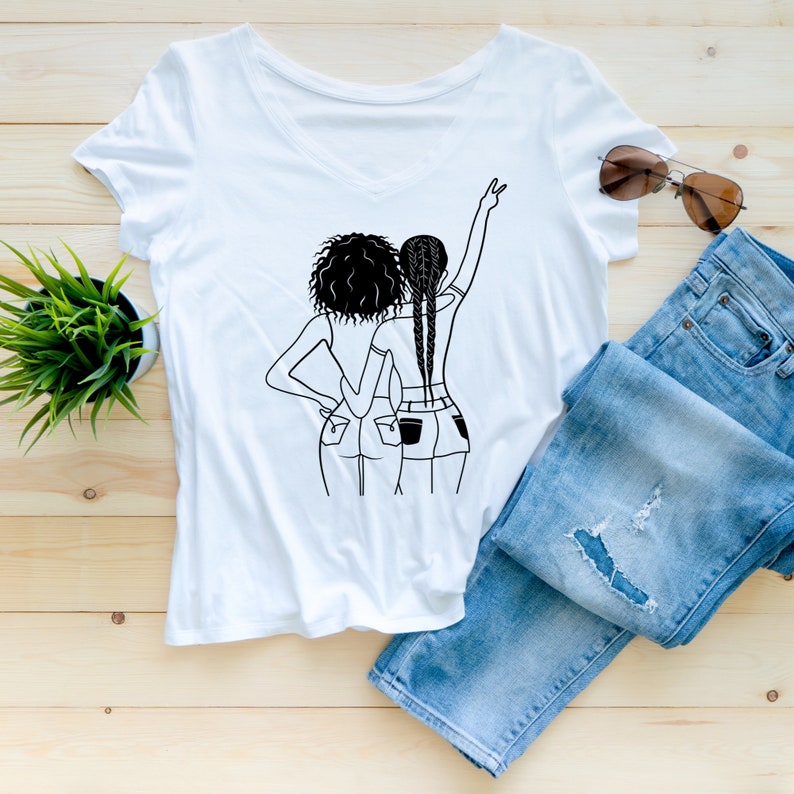May include: White t-shirt with a black line drawing of two women standing back to back, wearing jeans and one woman has her arm raised in the air.