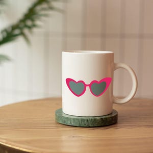 May include: White ceramic mug featuring a pink heart-shaped sunglasses design with gray lenses. The mug is sitting on a green coaster on a wooden surface. The mug has a curved handle.