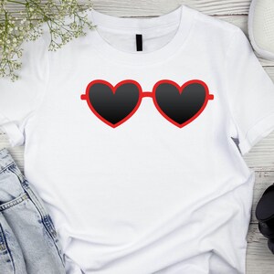 May include: White t-shirt featuring a graphic of heart-shaped sunglasses with black lenses and a red frame. The sunglasses are centered on the shirt. The shirt is on a light wood surface with a sprig of white flowers and a pair of jeans.