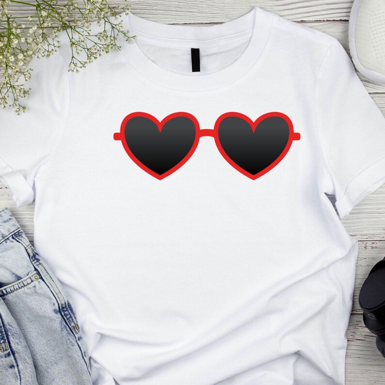 May include: White t-shirt featuring a graphic of heart-shaped sunglasses with black lenses and a red frame. The sunglasses are centered on the shirt. The shirt is on a light wood surface with a sprig of white flowers and a pair of jeans.