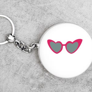 May include: A white keychain with a round charm featuring a pair of heart-shaped sunglasses in pink with gray lenses. The keychain has a silver chain and a silver key ring.