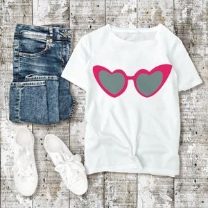 May include: A white t-shirt with a pink heart-shaped sunglasses graphic, paired with blue jeans and white sneakers. The sunglasses have gray lenses. The items are arranged on a weathered, light-colored wooden surface.