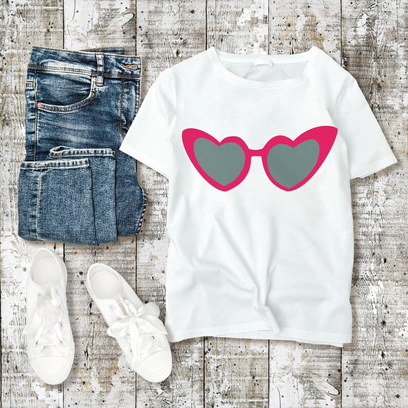 May include: A white t-shirt with a pink heart-shaped sunglasses graphic, paired with blue jeans and white sneakers. The sunglasses have gray lenses. The items are arranged on a weathered, light-colored wooden surface.