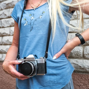 Op de afbeelding: Een blauw t-shirt met een grafische print van witte bloemen en gele bijen, gecombineerd met een zilveren en zwarte Praktica SLR-filmcamera die aan een zwarte leren nekriem hangt. De camera heeft een zwarte lensdop en een zwarte grip.