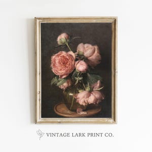 Op de afbeelding: Een vintage schilderij van een boeket roze pioenen in een glazen vaas. De bloemen zijn in een vaas op een houten tafel gerangschikt. Het schilderij is ingelijst in een lichtbruin frame.