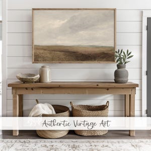 Op de afbeelding: Een rustieke houten consoletafel met twee geweven manden eronder. Boven de tafel hangt een ingelijst landschapsschilderij, met een keramische pot en een kom op de tafel. De muur achter de tafel is wit met horizontale houten lambrisering.