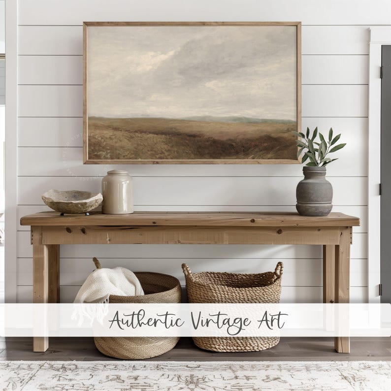 Op de afbeelding: Een rustieke houten consoletafel met twee geweven manden eronder. Boven de tafel hangt een ingelijst landschapsschilderij, met een keramische pot en een kom op de tafel. De muur achter de tafel is wit met horizontale houten lambrisering.