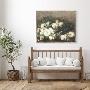 May include: A framed painting of white flowers with green leaves on a brown background. The painting is hanging on a white wall above a wooden bench with white cushions.