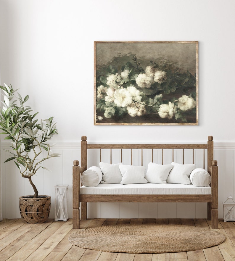 May include: A framed painting of white flowers with green leaves on a brown background. The painting is hanging on a white wall above a wooden bench with white cushions.