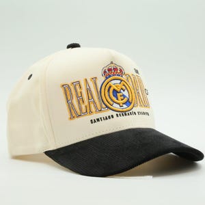 May include: Cream-colored baseball cap with a black corduroy brim. The cap features the Real Madrid logo in gold and blue, with the words "REAL MADRID" and "SANTIAGO BERNABEU STADIUM" embroidered on the front. A black button sits atop the cap.
