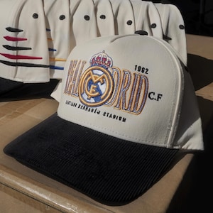 Puede incluir: Gorras de b&eacute;isbol color crema con viseras de pana negras. Una gorra presenta el texto "MADRID" con detalles en rojo, negro y azul. Otra gorra muestra el logo del Real Madrid, el texto "1902 C.F." y "SANTIAGO BERNAB&Eacute;U STADIUM".