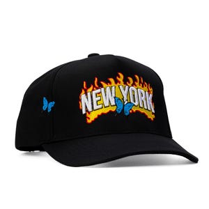 Può includere: Un cappellino da baseball nero con la scritta NEW YORK ricamata in bianco sul davanti, circondata da fiamme ricamate in giallo e arancione. Una piccola farfalla blu è ricamata sopra il testo e sul lato del cappellino.