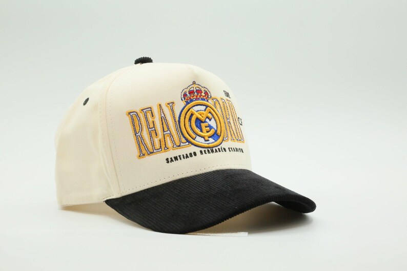 Puede incluir: Gorra de b&eacute;isbol color crema con visera de pana negra. La gorra presenta el logotipo del Real Madrid en dorado y azul, con las palabras "REAL MADRID" y "SANTIAGO BERNABEU STADIUM" bordadas en la parte delantera. Un bot&oacute;n negro en la parte superior.