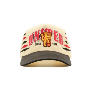 Op de afbeelding: Crèmekleurige baseballcap met een gebogen klep en een donkergrijze klep. De cap heeft het woord "UNITED" in het rood, een geel duivelslogo en de tekst "SINCE 1878". Zwarte en rode strepen zijn aan de zijkanten.