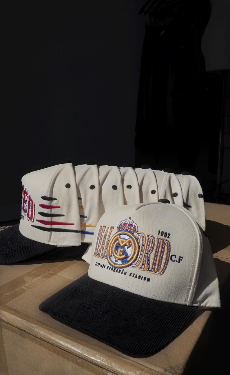 Puede incluir: Gorras de b&eacute;isbol color crema con viseras de pana negras. Una gorra presenta el texto "MADRID" con detalles en rojo, negro y azul. Otra gorra muestra el logo del Real Madrid, el texto "1902 C.F." y "SANTIAGO BERNAB&Eacute;U STADIUM".