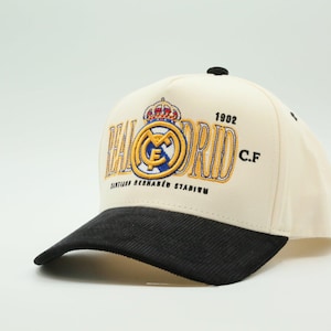 Puede incluir: Gorra de b&eacute;isbol color crema con visera negra. Presenta el logotipo del Real Madrid, el a&ntilde;o 1902 y el texto "SANTIAGO BERNAB&Eacute;U STADIUM" bordado en dorado y azul.