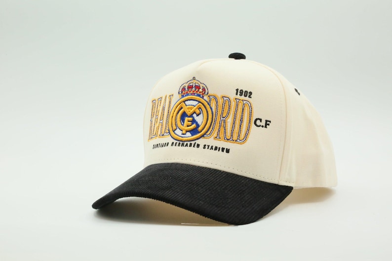 Puede incluir: Gorra de b&eacute;isbol color crema con visera negra. Presenta el logotipo del Real Madrid, el a&ntilde;o 1902 y el texto "SANTIAGO BERNAB&Eacute;U STADIUM" bordado en dorado y azul.