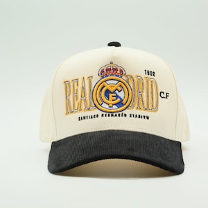 Puede incluir: Gorra de b&eacute;isbol color crema con visera de pana negra. La gorra presenta el logotipo y el texto del Real Madrid en dorado y azul, incluyendo "REAL MADRID C.F." y "1902". Tambi&eacute;n es visible el texto "SANTIAGO BERNAB&Eacute;U STADIUM".