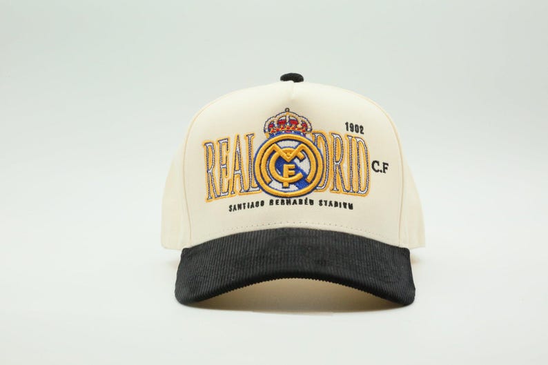 Puede incluir: Gorra de b&eacute;isbol color crema con visera de pana negra. La gorra presenta el logotipo y el texto del Real Madrid en dorado y azul, incluyendo "REAL MADRID C.F." y "1902". Tambi&eacute;n es visible el texto "SANTIAGO BERNAB&Eacute;U STADIUM".