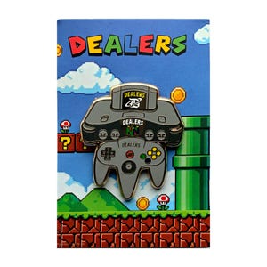May include: A grey enamel pin shaped like a video game console and controller, featuring the text DEALERS on the cartridge, console, and controller. The pin is displayed on a backing card with a blue sky, green pipe, and brick ground design.