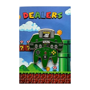 May include: A green enamel pin shaped like a video game console and controller, featuring the word DEALERS and a stylised logo. The pin is attached to a rectangular backing card with a blue sky, green hills, and brown brick patterns.