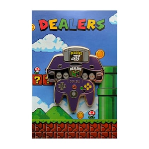May include: A purple enamel pin shaped like a video game console and controller, featuring the word DEALERS on the cartridge, console, and controller. The pin is displayed on a backing card with a pixelated landscape of green pipes and clouds.