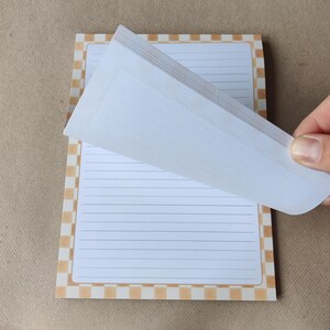 May include: A lined notepad with a brown and white checkered border. The top page is being lifted to reveal the pages underneath. The notepad is on a brown surface.