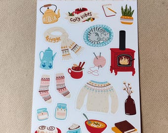 Op de afbeelding: Een stickervel met winterthema-illustraties. Ontwerpen omvatten een theepot, sjaal, kat, open haard, trui, sokken en andere gezellige items. De tekst "Cozy Winter" staat bovenaan het vel.