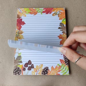 Può includere: Un blocco note a righe con un bordo a tema autunnale con foglie colorate, ghiande e funghi. La pagina superiore viene sollevata per rivelare le pagine sottostanti.
