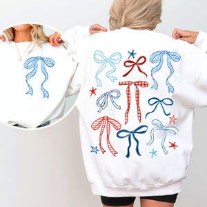 Puede incluir: Sudadera blanca con ilustraciones de lazos en azul y rojo, con motivos de estrellas. La parte delantera muestra un solo lazo azul. Los lazos tienen diferentes patrones, incluyendo lunares y cuadros. Una prenda casual y cómoda.