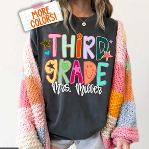 May include: A charcoal gray t-shirt featuring the colorful text "THIRD GRADE" in a playful, multi-colored font with star, flower, and smiley face accents. Below the text, the name "Mrs. Miller" is written in white cursive script.
