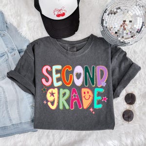 Puede incluir: Una camiseta gris oscuro con las palabras "SECOND GRADE" en letras coloridas y de burbujas. El dise&ntilde;o incluye estrellas, corazones y una cara sonriente. Se ve una gorra de camionero en blanco y negro con un dise&ntilde;o de cerezas.