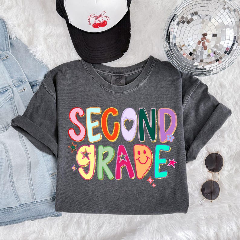 Puede incluir: Una camiseta gris oscuro con las palabras "SECOND GRADE" en letras coloridas y de burbujas. El dise&ntilde;o incluye estrellas, corazones y una cara sonriente. Se ve una gorra de camionero en blanco y negro con un dise&ntilde;o de cerezas.