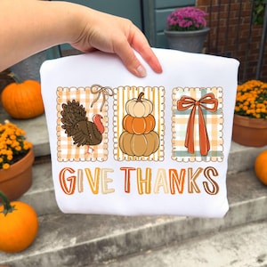 May include: White sweatshirt with a Thanksgiving-themed design. The design includes a turkey, stacked pumpkins, and a bow, all within decorative frames. The words "GIVE THANKS" are printed in orange and brown.