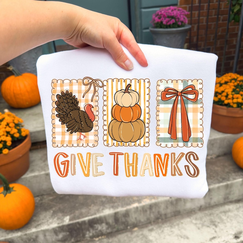 May include: White sweatshirt with a Thanksgiving-themed design. The design includes a turkey, stacked pumpkins, and a bow, all within decorative frames. The words "GIVE THANKS" are printed in orange and brown.