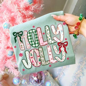 May include: A light green long-sleeve shirt with the words "HOLLY JOLLY" in a colorful, patterned design. The letters are filled with Christmas-themed patterns, including gingerbread men, trees, and bows. The shirt is held up by a hand.
