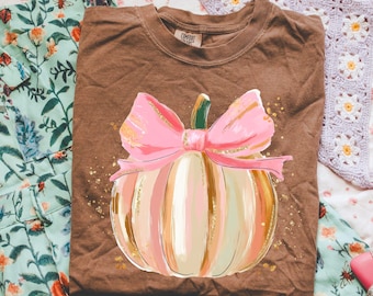 May include: A brown t-shirt featuring a colorful pumpkin design with a pink bow and gold accents. The pumpkin has vertical stripes in shades of pink, peach, and cream. The shirt is folded and lies on a floral patterned fabric.