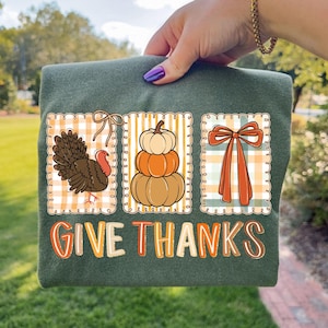 May include: Green t-shirt featuring three fall-themed illustrations: a turkey, stacked pumpkins, and a bow, set against a plaid background. The text "GIVE THANKS" is printed in a colorful, stylized font. The shirt is held up by a person.