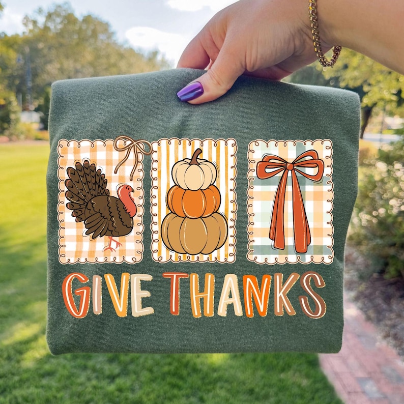 May include: Green t-shirt featuring three fall-themed illustrations: a turkey, stacked pumpkins, and a bow, set against a plaid background. The text "GIVE THANKS" is printed in a colorful, stylized font. The shirt is held up by a person.