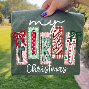 May include: A dark green long-sleeve shirt with the words "my FIRST Christmas" in white script. The letters are in colorful, festive designs with bows and Christmas tree accents, perfect for the holiday season.