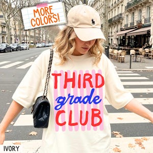 May include: An ivory t-shirt featuring the text THIRD grade CLUB in red and blue, layered over vertical pink stripes. The shirt is paired with a beige baseball cap with a small dark logo and a black quilted shoulder bag with a chain strap.
